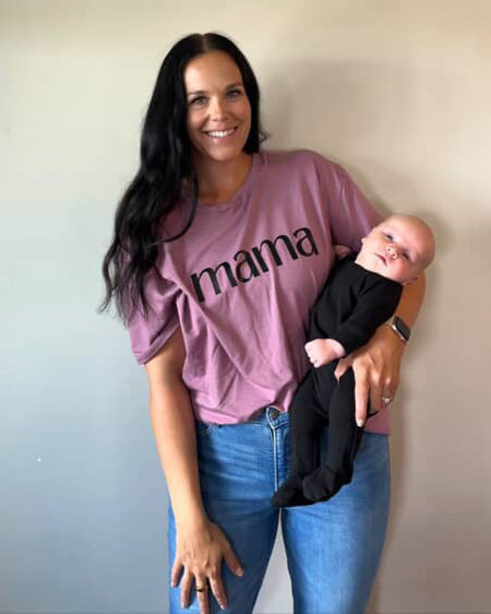 Woman wearing a 'mama' shirt holding a baby against a plain wall.