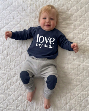 Baby lying on a white quilt in a navy love my dada sweater