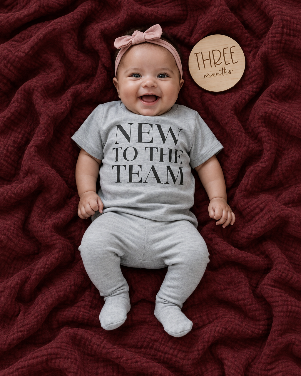 Baby on a burgundy blanket wearing a naturally draped NEW TO THE TEAM tee