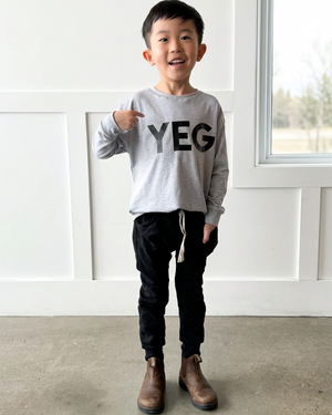 Boy in a long-sleeved grey YEG shirt and brown Chelsea boots, beside a window in a clean interior