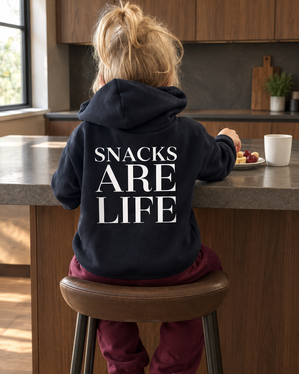 Girl having a snack at a moody kitchen island wearing a Snacks Are Life hoodie and burgundy pants