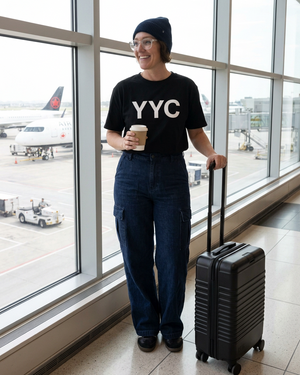 Woman standing and smiling to the side at the airport, holding coffee and black rolling luggage, wearing her YYC shirt, navy toque, and navy cargo jeans